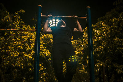 Man practicing chin-ups on gymnastic bars against trees at public park during night