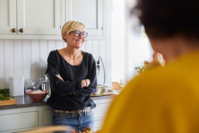 Smiling mature woman with arms crossed looking away in kitchen with friend at home
