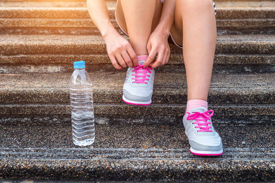 Low section of woman tying shoelace while sitting with water bottle on steps during exercising