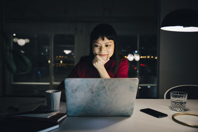 Confident businesswoman looking at laptop while sitting with hand on chin working late in office