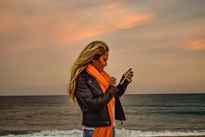 Young woman using phone while standing on beach against sky during sunset