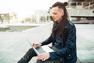 Hipster man using laptop on footpath in city