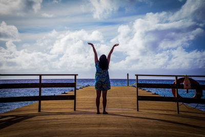 Rear view of woman standing on pier with arms raised against sky