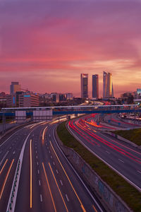 Light trails on road by illuminated buildings against sky during sunset