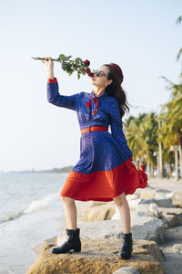 Woman holding rose while standing on beach against sky
