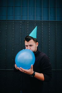 Man wearing party hat blowing balloon against metal grate