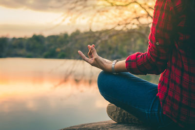 Cropped image of young woman meditating while sitting on rock by lake during sunset