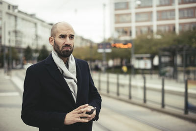 Thoughtful mid adult businessman holding smart phone on sidewalk in city