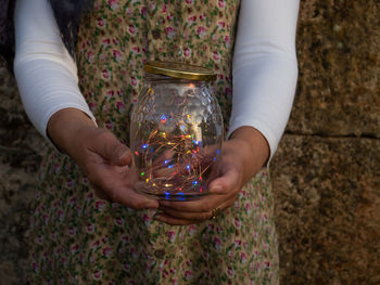 Midsection of woman holding string lights in jar against stone wall