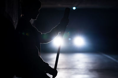Side view of contemporary female in bomber jacket holding a black baseball bat on shoulder with car lights on background