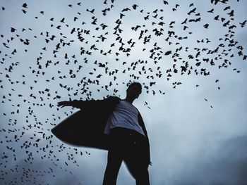 Low angle view of man standing under silhouette birds flying against sky