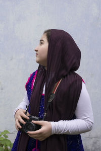 Girl with camera looking away while standing against wall