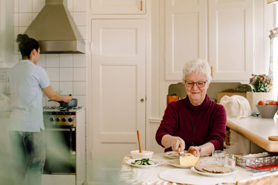 Smiling retired elderly woman eating breakfast while female caregiver preparing food on stove in kitchen at nursing home