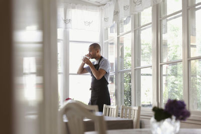 Male owner having coffee and talking on phone while standing by window at cafe