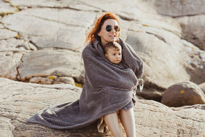 Mother and baby girl wrapped in blanket while sitting on rock formation during summer