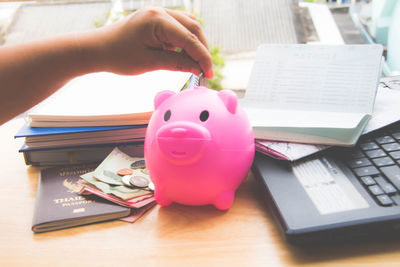 Cropped hand of person putting coin in piggybank by passport with books on table