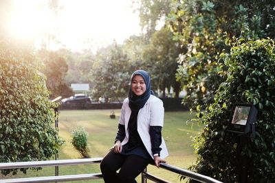 Portrait of smiling young woman wearing hijab sitting on railing against trees in park