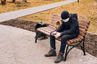 Boy wearing mask using phone sitting on bench at park
