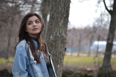 Thoughtful young woman wearing denim jacket against bare trees