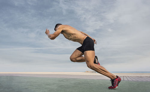 Side view of shirtless man taking running stance against sky