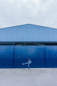 Cheerful remote view of female in denim apparel jumping along street on background of blue building