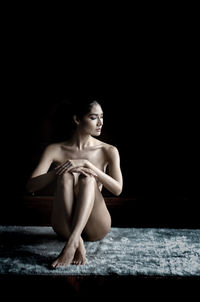 Naked young female model sitting on rug against black background