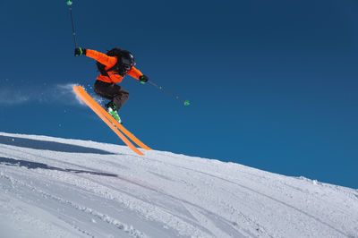 A skier in an orange jacket and a black helmet makes a jump on a sunny day against a clear sky. copy