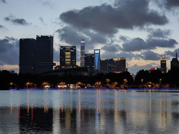 Illuminated buildings by river against cloudy sky