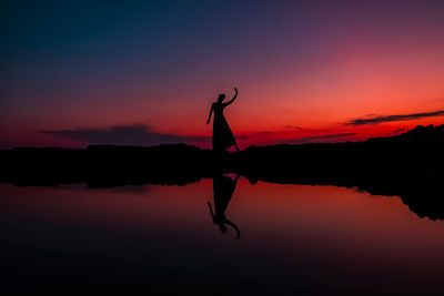 Rear view of silhouette woman standing by lake against sky during sunset