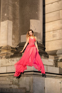 Full length of young woman in pink evening gown sitting against columns