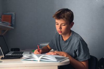 Student teenager boy doing homework with laptop, open copybook and computer, workplace at home