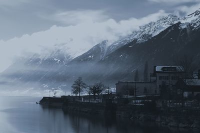 Buildings by lake against mountains during foggy weather