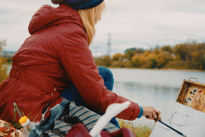 Side view of woman painting while sitting at lakeshore