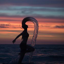 Side view of silhouette woman tossing wet hair in sea against sky during sunset