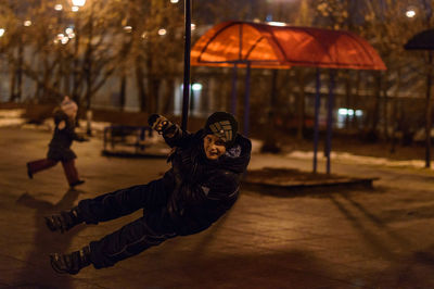 Full length portrait of boy swinging in city at night during winter