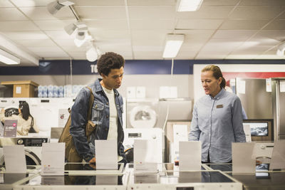 Mature female owner explaining about appliance to young customer while standing in electronics store