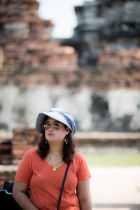 Portrait of beautiful young woman wearing hat standing against built structure