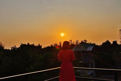 Rear view of man standing by railing against sky during sunset