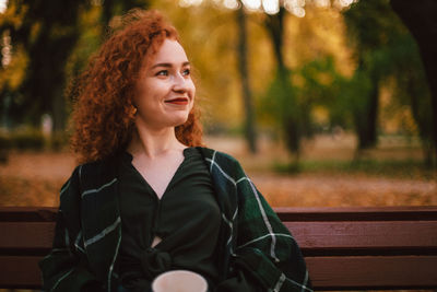 Portrait of smiling redhead woman sitting on bench in park in autumn