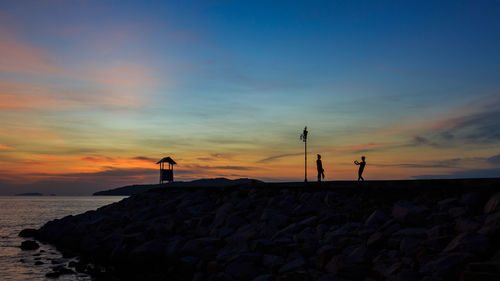 Silhouette friends standing on rock by sea against sky during sunset