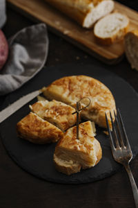 Slice of spanish potato omelette served on a bread slice and a silver old fork. shot taken from high angle with bread in a wooden cutting board in the background.