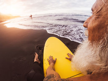 Close-up of senior man with surfboard sitting at beach