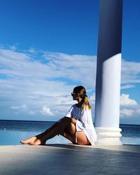 Young woman sitting by column while looking at sea against sky