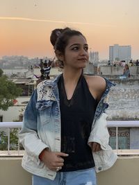 Portrait of young woman standing against cityscape during sunset