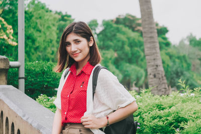 Portrait of smiling woman standing by railing against trees