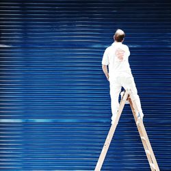 Rear view of man standing on ladder against blue corrugated iron