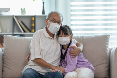 Portrait of grandfather and granddaughter wearing mask sitting on sofa at home