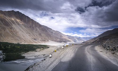 Empty road by majestic mountains against cloudy sky