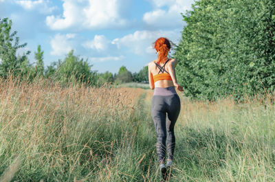 A young female runner runs along the road overgrown with grass. rear view