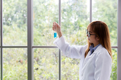 Scientist looking at test tube with blue liquid while standing against window